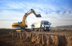 A huge yellow crawler excavator and a construction dump truck standing next to it while working on a sunny day against a blue sky. Excavator digs a pit and loads a dump truck with earth