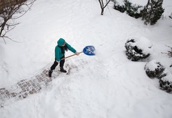 Unrecognizable young woman in a hood, clearing the sidewalk, street from snow after a strong blizzard, holding a snow shovel. Winter season, bad weather, cold, snowy