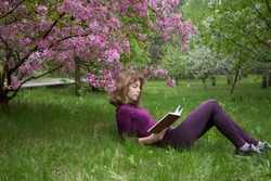 young woman, student, reads book with interest, lying under tree on spring day in park. girls - teenager 17 years old with book on grass. preparation for exams. Spring leisure. Passion for reading