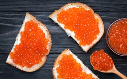 Salmon Red Caviar. Red fish caviar on a spoon in glass bowl. Sandwich with white bread, butter and red caviar. Raw seafood. Luxury delicacy food. Dark natural wooden background. Flat lay, top view.