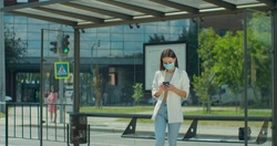 Front view of young woman using smartphone while waiting at public transport stop. Girl in medical face mask looking at phone screen while browsing internet. Health and safety, quarantine.