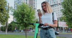 Pretty woman is using smartphone after ride on electric scooter in the urban city. Woman texts sms using smartphone near electric scooter.