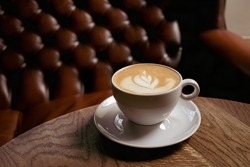 Atmospheric cup of cappuccino with latte art on the wooden table and brown leather armchair as background. Cafe and bar, barista art concept. Soft focus. Coffee and relax time. Dark cozy composition.