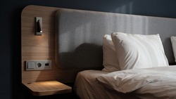 Close-up fragment of bedroom with empty bedside table, reading lamp and a USB socket in modern interior​ design home or hotel. Soft pillow and blanket, stylish comfortable furniture. Sun shadows.