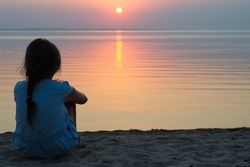 girl sitting on the beach in a light summer dress, watching the sun set into the sea on the horizon