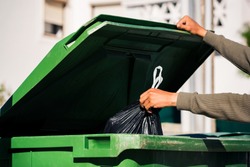 Man throwing out black eco-friendly recyclable trash bag in to big plastic green garbage container. Take out the trash