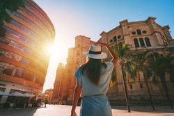 Rear view of young tourist woman in white sun hat walking in Malaga city at sunset. Summer holiday vacation in Spain