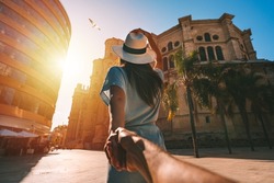 Follow me - POV. Young tourist woman in white sun hat holding her boyfriend by hand and walking in Malaga city at sunset. Couple on summer holiday vacation in Spain. Traveling together.