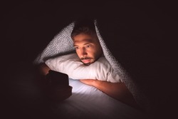 Young man lying in bed under a blanket and using smartphone at night. Social media addiction and communication