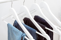 Many blue denim jeans hanging on white clothes hangers on clothing rack. Close up of folded casual denim jeans in wardrobe with coat hangers