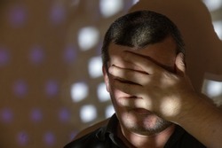 A man covered his face with his hand. Portrait of a middle-aged man against a wall. A light and shadow pattern of repeating figures on the wall and the man's face. People, shame, disgrace concept