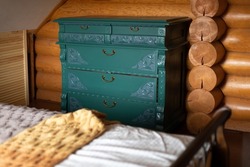Old renovated chest of drawers with carved patterns painted turquoise located by wall behind double bed in wooden country house. Eco-friendly re-use - recycling . Environmental friendliness. 