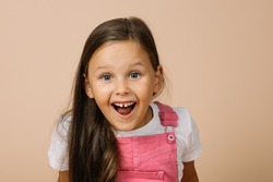 Little girl with surprised eyes, opened mouth, blissful smile with teeth and raised eyebrows looking at camera wearing bright pink jumpsuit and white t-shirt on beige background.