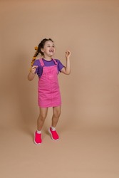 Delighted thrilled jumping small child with yellow kanekalon pigtails dancing looking up smiling with missing tooth in pink jumpsuit, purple t-shirt and pink sneakers on beige background.