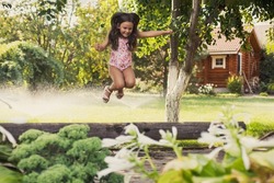 Excited female child playing outside jumping off log looking down in backyard indulging with trees and water sprinklers in background in daytime. Dreaming to become sports gymnast.