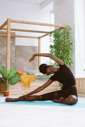 Smiling, glad flexible sportive barefoot dark skin interracial woman stretching, doing yoga, fitness exercises on blue mat at home. Training for health indoors. Side bend stretching, body incline.