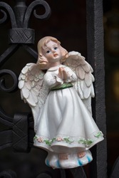 Figurine of an angel in a white dress. Decorative figure on a metal railings background. Statuette. Macro photography of isolated figurine of an angel hanging on the railings..