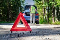 A portable, reflective red warning sign stands on the side of the road next to the damaged vehicle. A driver in a yellow vest is standing near the trunk. Selective focus.