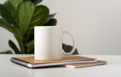 Mockup of a white mug on a table in a minimalist interior, business notepads, Ficus Lyrata Compacta. Template, layout for your design, advertising, logo with copy space. Cup on light gray background.