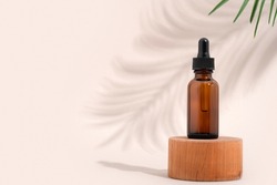 Mockup cosmetic face serum brown glass bottle with a pipette on a stylish minimalist beige white background wooden pedestal. Shadow from a palm tree branch. Female beauty concept, empty template