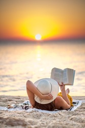 Young woman reading a book at the beach while the sun rise. She's wearing a orange dress and white hat