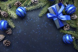 Christmas gift with blue ribbon and blue balls, tree branches and cones on dark blue background with copy space.