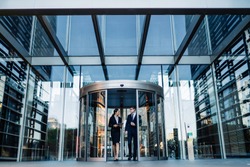 Formal contemporary man and woman talking while walking out of round revolving doors of modern glass building of corporation office 