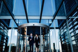 Formal multiracial coworking woman and man in suits walking out of glass round revolving doors of office building and talking 