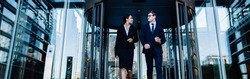 Low angle of young smiling multiethnic woman and man with coffee cup walking out of glass revolving doorway of glass building
