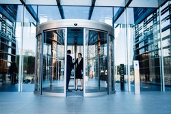Contemporary diverse formal man and woman standing together inside of round revolving doors of modern glass office building and talking 