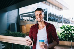 Half length portrait of cheerful hipster guy in casual clothes smiling at camera during leisure time with coffee, happy Caucaisan blogger with takeaway cup and modern cellphone device posing
