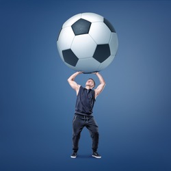 A muscular man tries to hold a giant and heavy black and white football ball over himself. Football and soccer. Sport gear. Football champion.