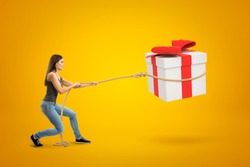 Side view of young woman standing with bent knees and pulling big gift box in air which she has lassoed, on yellow background. Bargain bin. Good eye for bargain. Get what you want.