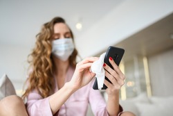 European woman in face mask cleaning the phone by hand sanitizer, using cotton wool with alcohol to wipe to avoid contaminating with Corona virus. Cleaning mobile phone to eliminate germs, Covid-19. 