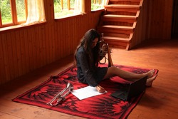 Portrait of a happy young woman with musical instrument and laptop.Girl with long hair composing music on a white sheet sitting on the floor holding a trumpet with bare legs in a wooden country house