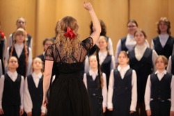 Woman singer blonde in black dress with raised hand leads the choir standing in front of a group of children students of a music school back view of the leader