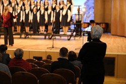A person with a camera at a concert shoots a group of children with a conductor singing standing in a row on stage