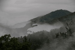 Terrible huge tyrannosaurus in the fog climbs the slope of a mountain covered with green forest