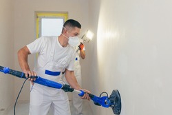 Plasterer smoothes the wall surface with a wall grinder. Two master builders grind a white plaster wall. a man in overalls grinds the surface in a respirator. experienced repairman
