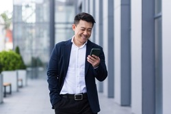 asian male freelancer walking near business center holding phone, smiling reading news, successful businessman