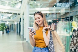 Young woman consumer in the mall browses chat and uses using a smartphone. female standing with a mobile phone in her hands in shopping center. indoor. happy shopper girl with gift bags make purchases