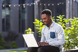 black man freelancer working online using laptop sitting on bench outside office modern building in city urban park on street. Happy African american guy student in casual outdoors typing on keyboard