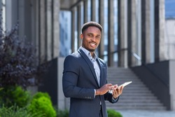 Young african american businessman in formal business suit standing working with tablet in hands on background modern office building outside. Man using smartphone or mobile phone outdoors city street