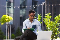 Young african american male student sitting in a city park on a bench with laptop and notebook studying online outdoors Man freelancer in glasses with books learn working remotely in street e-learning