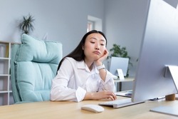 Boring and monotonous work. A young beautiful Asian business woman, an office worker sits at a desk at a computer, is sad, bored, performs monotonous work. He holds his head with one hand.