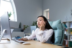 Boring and monotonous work. A young beautiful Asian business woman, an office worker sits at a desk at a computer, is sad, bored, performs monotonous work. He holds his head with one hand.