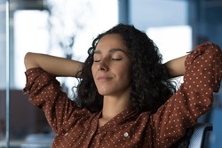 Close-up photo portrait of young beautiful curly Arab woman resting indoors at home, woman with hands behind head resting sitting on sofa near window, eyes closed breathing, meditating and dreaming