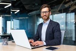 Portrait of mature boss inside office with laptop, successful and satisfied investor manager looking at camera and smiling man in glasses and business suit, investor with beard sitting on chair.