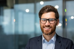 Close up photo portrait of successful and happy businessman, mature boss with beard and glasses working inside modern office building, senior investor smiling and looking at camera