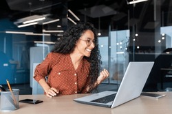Hispanic business woman celebrating victory success, employee with curly hair inside office reading good news, using laptop at work inside office holding hand up and happy triumph gesture.